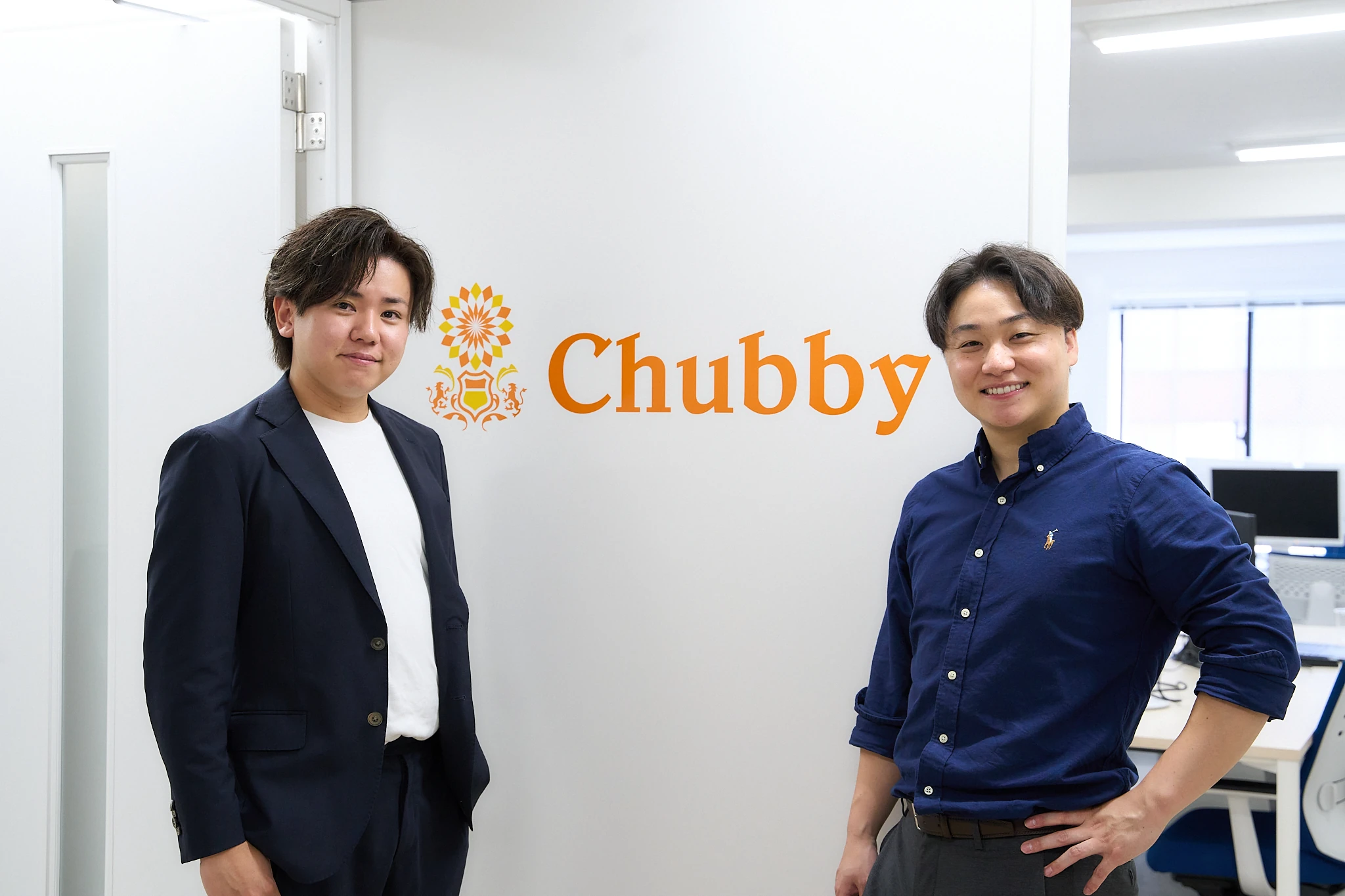 Chubbyのロゴの前に立つ代表取締役2名